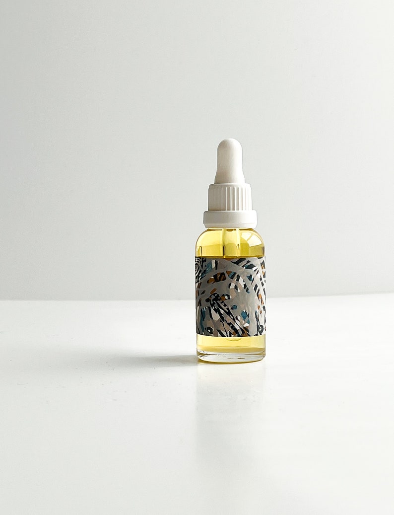 May include: A clear glass bottle with a white dropper top, filled with a yellow liquid. The bottle has a decorative label with a pattern of leaves in shades of gray, blue, and brown. The bottle is sitting on a white surface.