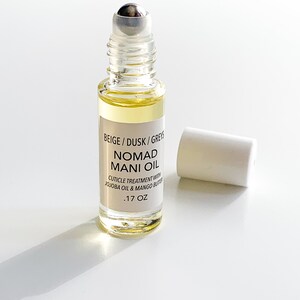 Puede incluir: Un frasco de vidrio transparente con Nomad Mani Oil, un tratamiento para cut&iacute;culas. El frasco contiene un l&iacute;quido amarillo claro y tiene un aplicador de bola plateada. La etiqueta dice "BEIGE / DUSK / GREYS NOMAD MANI OIL" y "CUTICLE TREATMENT WITH JOJOBA OIL & MANGO BUTTER .17 OZ". La tapa blanca est&aacute; quitada.