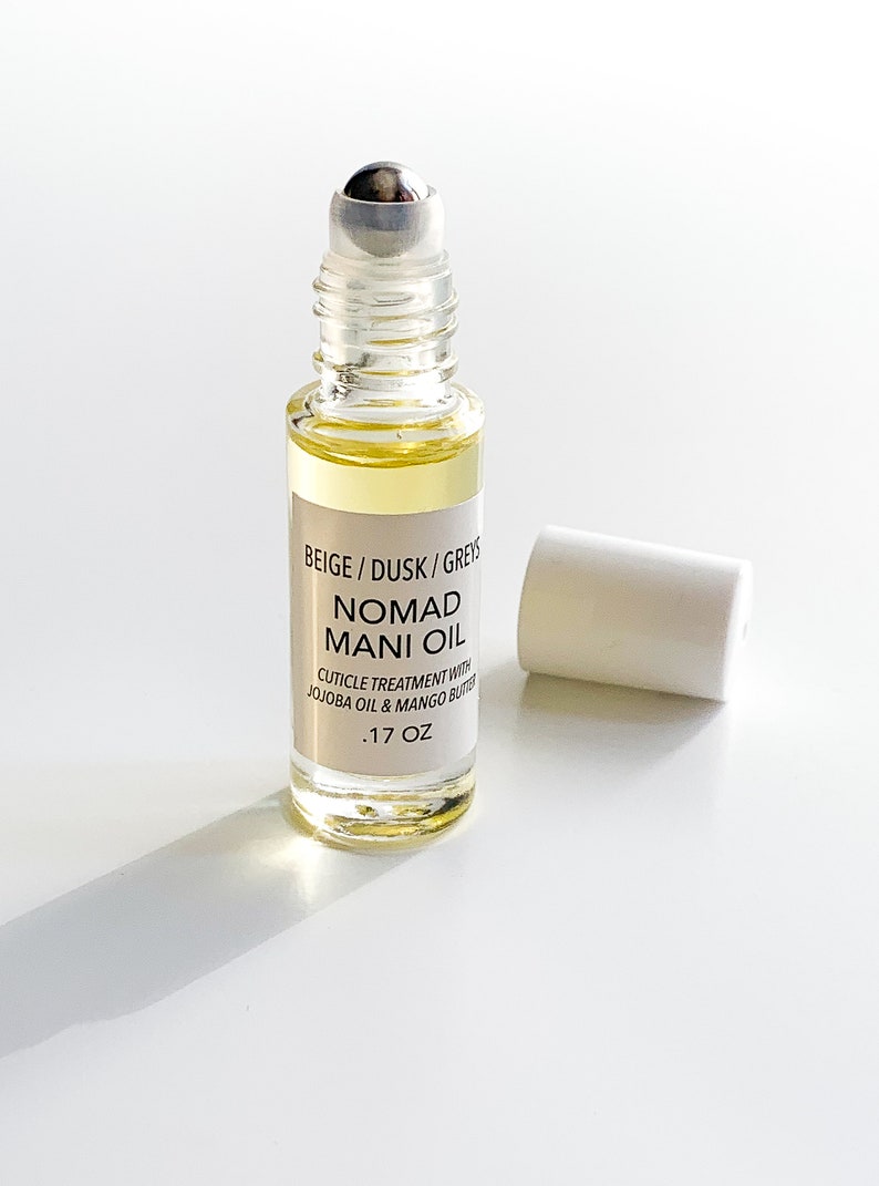 Puede incluir: Un frasco de vidrio transparente con Nomad Mani Oil, un tratamiento para cut&iacute;culas. El frasco contiene un l&iacute;quido amarillo claro y tiene un aplicador de bola plateada. La etiqueta dice "BEIGE / DUSK / GREYS NOMAD MANI OIL" y "CUTICLE TREATMENT WITH JOJOBA OIL & MANGO BUTTER .17 OZ". La tapa blanca est&aacute; quitada.