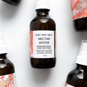 May include: Several amber glass bottles of Nectar Water Nourishing Rose & Honey Face Toner are arranged on a white surface. Each bottle has a white spray nozzle and a label with text. The labels have a beige and coral design. The bottles contain 3 oz of product.