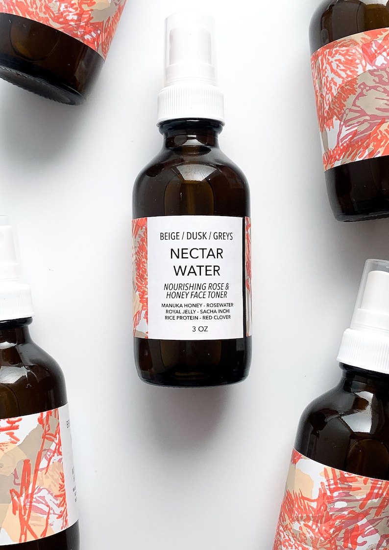 May include: Several amber glass bottles of Nectar Water Nourishing Rose & Honey Face Toner are arranged on a white surface. Each bottle has a white spray nozzle and a label with text. The labels have a beige and coral design. The bottles contain 3 oz of product.