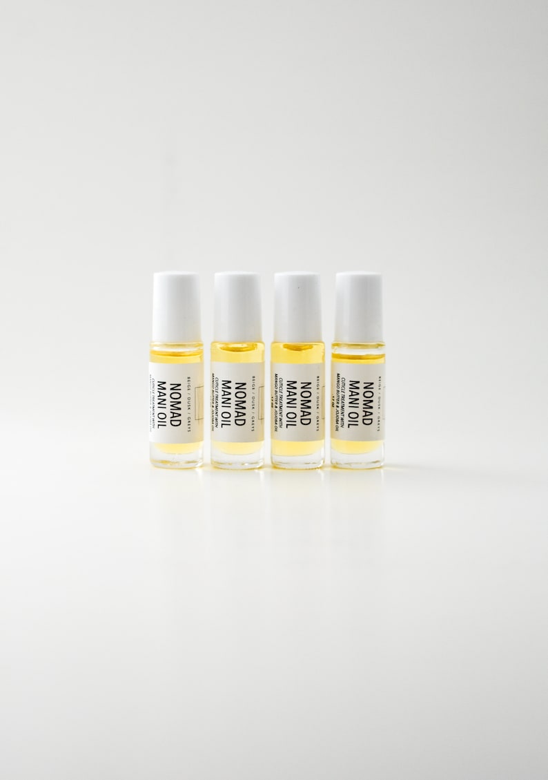 Puede incluir: Cuatro peque&ntilde;os frascos roll-on de vidrio transparente con tapas blancas est&aacute;n alineados. Cada frasco contiene un l&iacute;quido amarillo y tiene una etiqueta que dice "NOMAD MANI OIL". Las etiquetas tambi&eacute;n incluyen texto sobre el tratamiento de cut&iacute;culas.
