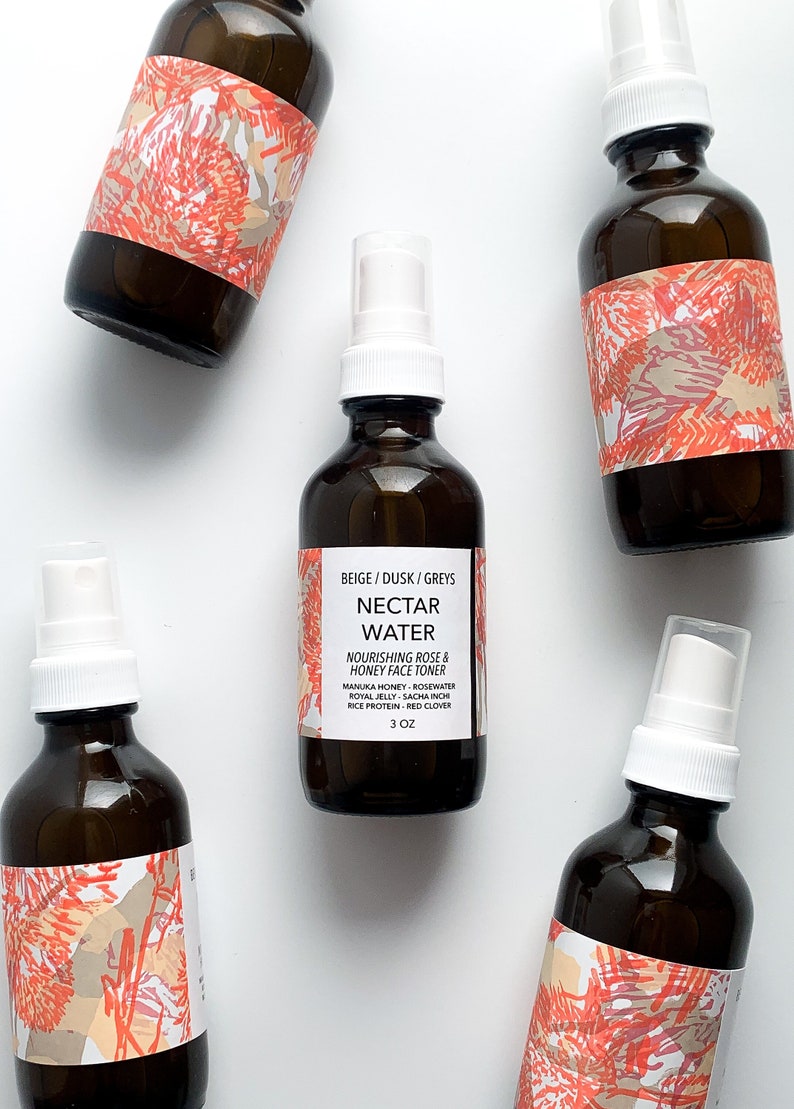 May include: Several amber glass bottles with white spray nozzles are arranged on a white surface. The bottles have labels with the text "Nectar Water" and "Nourishing Rose & Honey Face Toner." Each bottle has a decorative label with a coral and beige design.