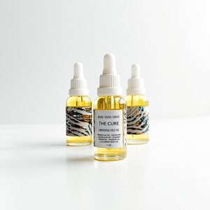 May include: Three glass bottles of face oil with white droppers. The bottles are filled with a yellow liquid and have labels with the text "THE CURE UNIVERSAL FACE OIL". The labels also list ingredients and the size, 1 oz.