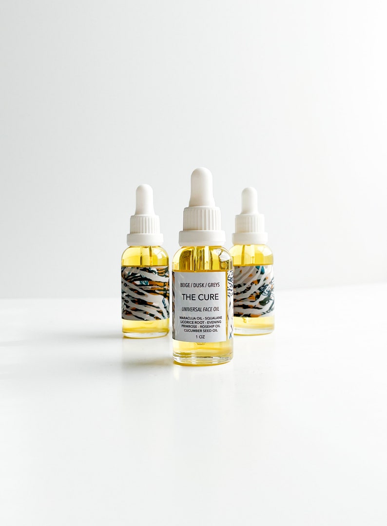 May include: Three glass bottles of face oil with white droppers. The bottles are filled with a yellow liquid and have labels with the text "THE CURE UNIVERSAL FACE OIL". The labels also list ingredients and the size, 1 oz.