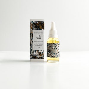 May include: A clear glass bottle with a dropper and a matching box. The bottle contains a yellow liquid and is labeled "THE CURE UNIVERSAL FACE OIL." The box and bottle have a beige, brown, and gray pattern. The bottle is 1 oz.