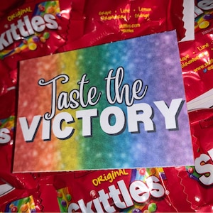 Könnte beinhalten: Eine Karte in Regenbogenfarben mit dem Text "Taste the VICTORY" darauf. Die Karte ist von Tüten mit Skittles-Bonbons umgeben.