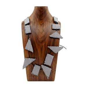 Puede incluir: Un collar que presenta múltiples formas de fieltro gris claro, incluyendo rectángulos y triángulos, conectados por un cordón negro fino. Cada pieza geométrica está terminada con costuras oscuras visibles a lo largo de los bordes, creando un aspecto estructurado.