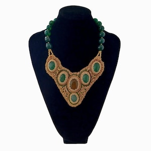 Può includere: Una collana di perline con pietre ovali verdi e marroni e perline color oro. La collana è esposta su un manichino nero. La collana ha un design a pettorina a forma di V.