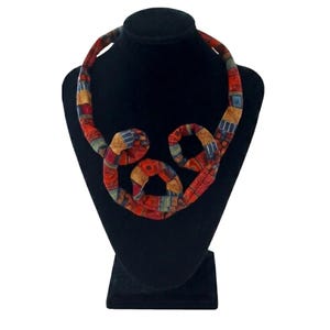 Könnte beinhalten: Ein farbenfrohes Stoffhalsband mit einem einzigartigen, abstrakten Design. Die Halskette besteht aus einer Mischung aus rot, orange, gelb und blau gemustertem Stoff, der auf einer schwarzen Schaufensterpuppe präsentiert wird. Die Halskette hat ein auffälliges Aussehen.