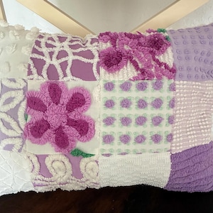 May include: A patchwork pillow with a variety of textures and patterns. The pillow features squares of white, lavender, and purple fabrics, including a large, textured flower in the center. The pillow is rectangular and has a soft, inviting appearance.