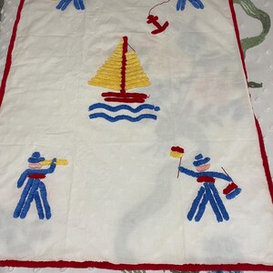 May include: A white baby blanket with a red border. The blanket features a yellow sailboat with blue waves, a red anchor, and four blue figures with red hats and accessories.