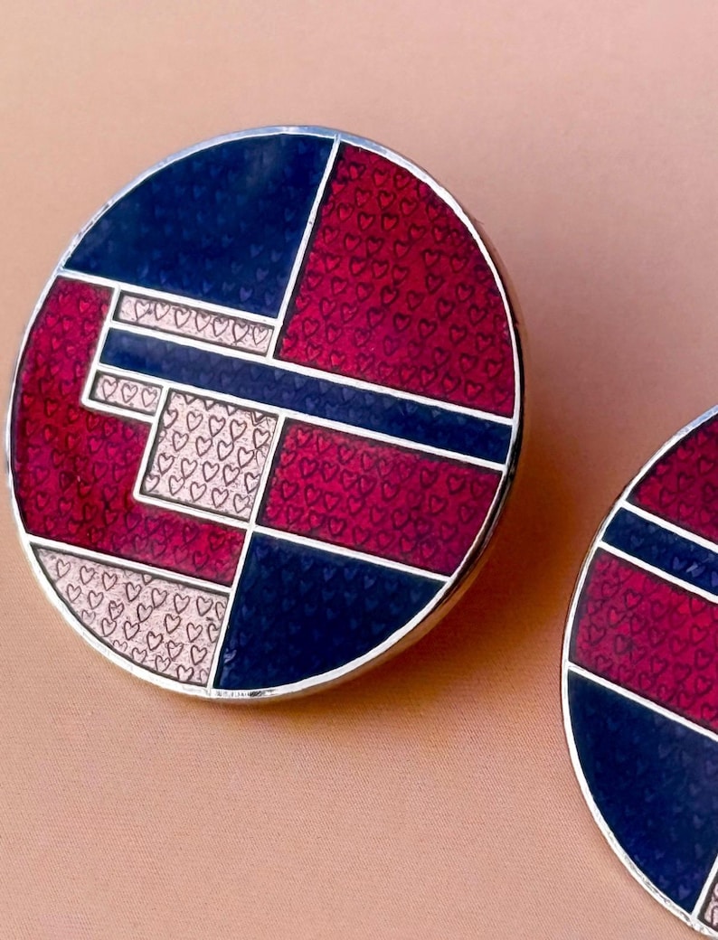 Peut inclure: Une paire de boucles d'oreilles rondes avec un motif g&eacute;om&eacute;trique. Les boucles d'oreilles pr&eacute;sentent une combinaison de sections bleu marine, bordeaux et rose clair. Les sections bordeaux sont recouvertes de minuscules c&oelig;urs, et les sections rose clair ont une bordure en forme de c&oelig;ur.