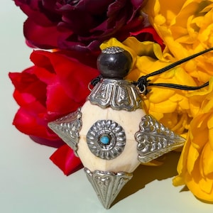 May include: A pendant necklace with a cream-colored, star-shaped pendant. The pendant has silver-colored metal accents and a small turquoise stone in the center. The necklace is set against a backdrop of red, yellow, and burgundy flowers.