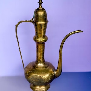 May include: A tall, polished brass ewer featuring an ornate, engraved floral pattern across its spherical base, slender neck, and domed lid. The vessel includes a long, curved spout and a thin, arched handle attached to the neck and body.