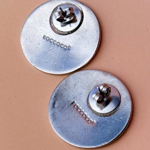 Peut inclure: Deux boucles d'oreilles rondes argent&eacute;es avec fermoirs &agrave; tige. Les boucles d'oreilles ont une surface textur&eacute;e et le mot "ROCCOCO" est estampill&eacute; au dos de chaque boucle d'oreille. Les boucles d'oreilles sont pr&eacute;sent&eacute;es sur une surface rose clair.