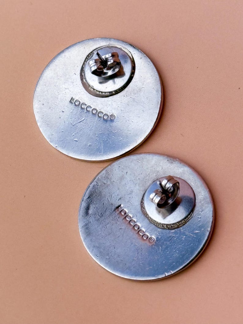 Peut inclure: Deux boucles d'oreilles rondes argent&eacute;es avec fermoirs &agrave; tige. Les boucles d'oreilles ont une surface textur&eacute;e et le mot "ROCCOCO" est estampill&eacute; au dos de chaque boucle d'oreille. Les boucles d'oreilles sont pr&eacute;sent&eacute;es sur une surface rose clair.