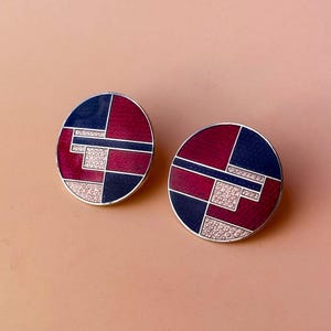 Peut inclure: Deux boucles d'oreilles rondes avec un motif g&eacute;om&eacute;trique. Chaque boucle d'oreille pr&eacute;sente une bordure argent&eacute;e, avec des sections bleu marine, bordeaux et une section avec de petits d&eacute;tails en forme de c&oelig;ur. Les boucles d'oreilles sont pos&eacute;es sur un fond rose clair.
