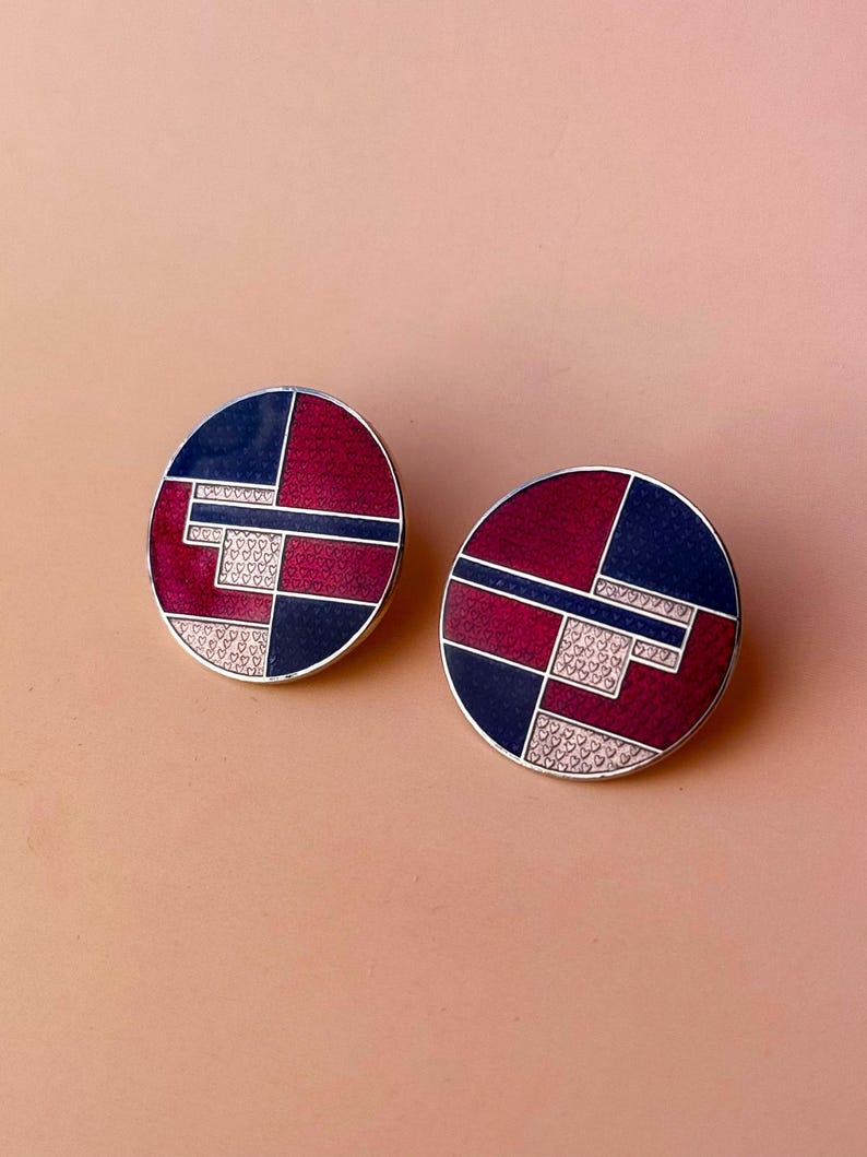 Peut inclure: Deux boucles d'oreilles rondes avec un motif g&eacute;om&eacute;trique. Chaque boucle d'oreille pr&eacute;sente une bordure argent&eacute;e, avec des sections bleu marine, bordeaux et une section avec de petits d&eacute;tails en forme de c&oelig;ur. Les boucles d'oreilles sont pos&eacute;es sur un fond rose clair.