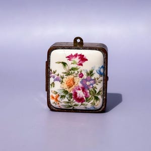 May include: A small, square trinket box with a floral design. The box features a white porcelain top with a colorful flower arrangement in shades of pink, purple, yellow, and blue. The box has a metal frame and a small loop at the top.