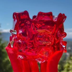 May include: A vibrant red glass bowl featuring a thick, textured surface with prominent raised circular indentations and a wavy, uneven rim. The translucent material catches the light, creating deep crimson highlights and shadows across the body.