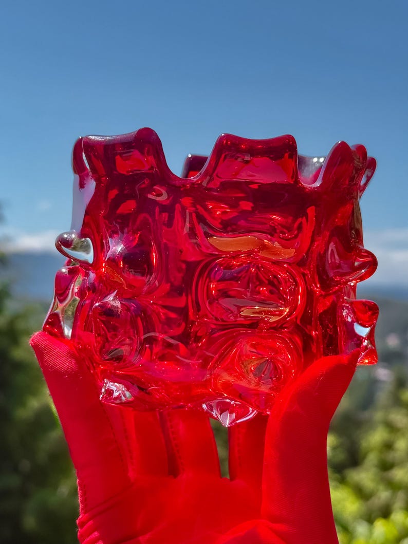 May include: A vibrant red glass bowl featuring a thick, textured surface with prominent raised circular indentations and a wavy, uneven rim. The translucent material catches the light, creating deep crimson highlights and shadows across the body.