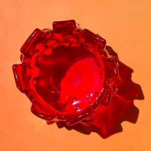 May include: A top-down view of a vibrant red glass bowl featuring a scalloped, petal-like rim. The translucent glass catches the light, creating a deep crimson hue and casting a distinct, sharp shadow on the surface beneath the object.