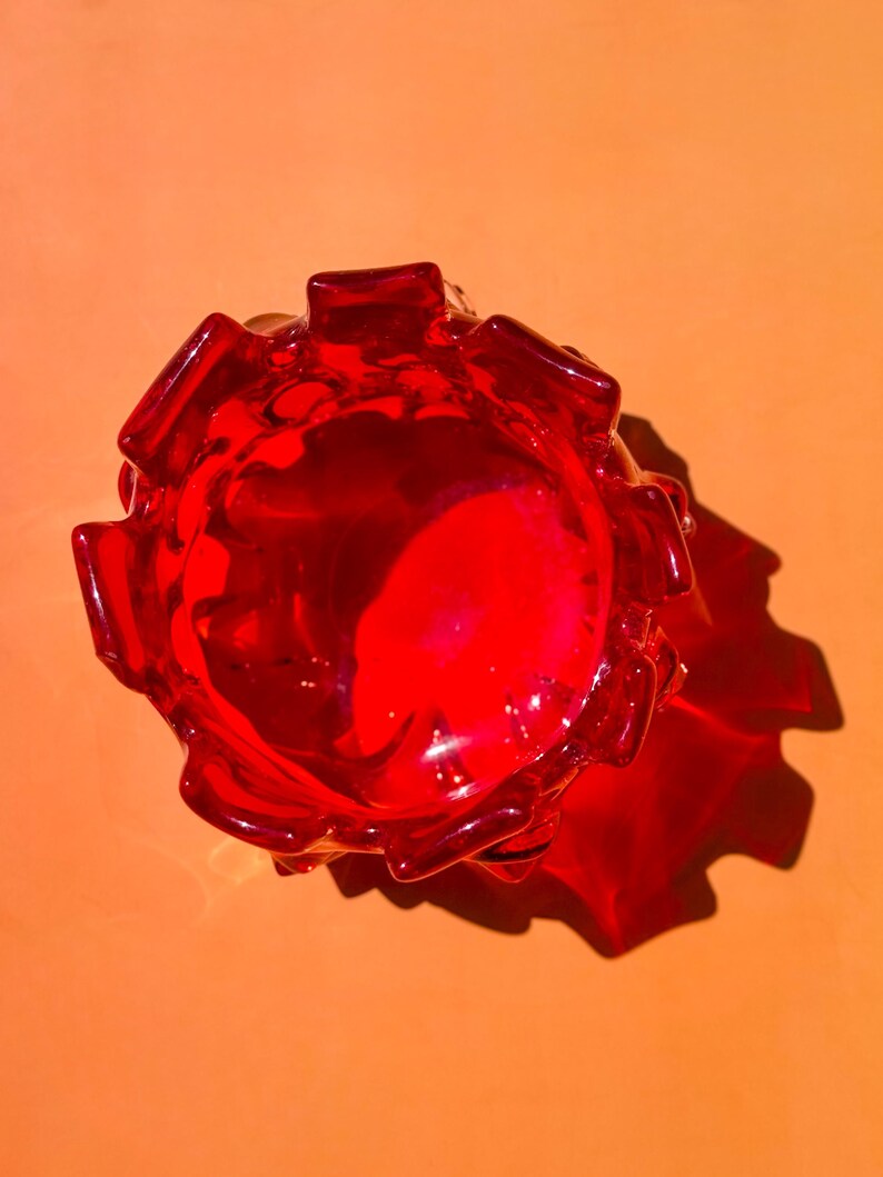 May include: A top-down view of a vibrant red glass bowl featuring a scalloped, petal-like rim. The translucent glass catches the light, creating a deep crimson hue and casting a distinct, sharp shadow on the surface beneath the object.