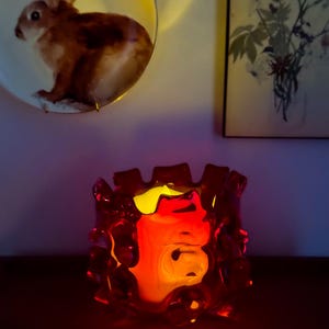 May include: A deep red glass candle holder with a thick, irregular, and textured rim. The vessel glows from within with a warm, bright yellow light, casting a soft illumination that highlights the translucent, organic shape of the glass structure.