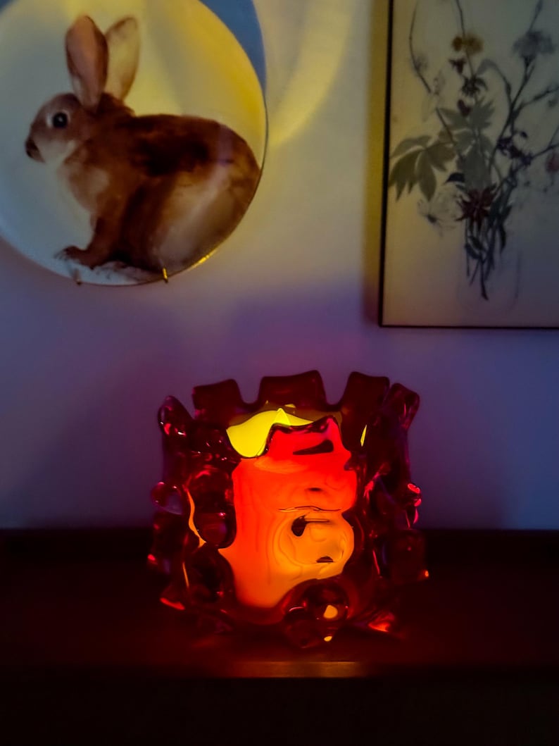 May include: A deep red glass candle holder with a thick, irregular, and textured rim. The vessel glows from within with a warm, bright yellow light, casting a soft illumination that highlights the translucent, organic shape of the glass structure.