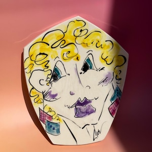 May include: A pentagonal ceramic wall plaque featuring a stylized line drawing of a face with curly yellow hair, purple lips, and geometric earrings in blue and pink. The artwork includes black ink outlines and a signature in the bottom right corner.