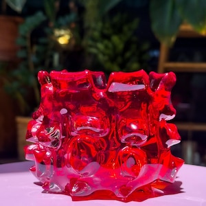 May include: A vibrant red glass votive holder featuring a thick, irregular, and textured surface with deep indentations and raised protrusions. The translucent material catches light, creating varied shades of red across its organic, sculptural form.
