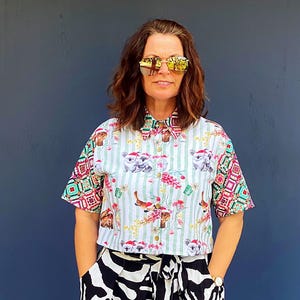May include: A short-sleeved button-down shirt featuring a light blue striped pattern with koala and holiday-themed graphics, paired with wide-leg trousers displaying a bold black and white zebra print, a tie-waist belt, and white trainers.