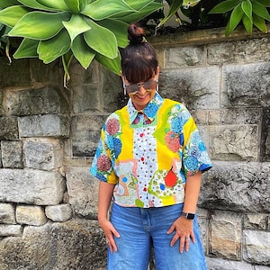 May include: A woman in a colourful, short-sleeved shirt with a patchwork design, paired with wide-leg blue jeans and silver sandals. The shirt features yellow, blue, and white patterns. A stone wall and large green plants form the background.