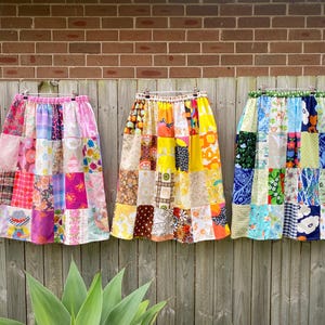 Può includere: Tre gonne patchwork colorate appese a una staccionata di legno. Le gonne presentano una varietà di motivi e colori di tessuto, tra cui rosa, giallo, blu e verde. Ogni gonna è composta da più quadrati di tessuto cuciti insieme.