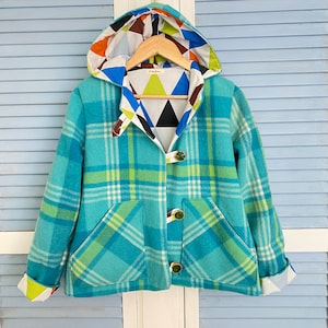 May include: A turquoise plaid hooded jacket with a colourful geometric patterned lining and hood. The jacket has toggle closures and two front pockets. The cuffs are also lined with a geometric pattern. The jacket is displayed on a wooden hanger.