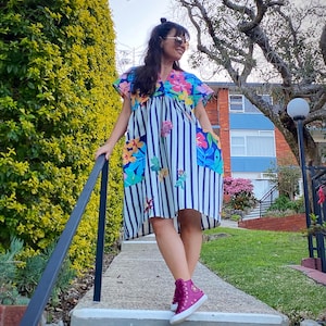 May include: A short-sleeved dress with a blue and white striped pattern and floral accents. The dress features a V-neckline and pockets with floral designs. The person is wearing pink high-top trainers.