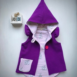 Könnte beinhalten: Eine lila Kapuzenweste für Kinder. Die Weste hat eine spitze Kapuze, einen rosa Knopfverschluss und eine Tasche. Das Innenfutter ist aus hellrosa Stoff mit einem Hasenmuster. Die West hat eine verspielte, märchenhafte Ästhetik.