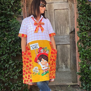 Könnte beinhalten: Vintage-Kleid mit weiß-orange kariertem Oberteil und Schleife. Die gelbe Schürze zeigt einen Cartoon und den Text "WOMEN'S LIB." Das Kleid hat orangefarbene Blumentaschen und -besätze. Die Person trägt schwarze Sandalen.