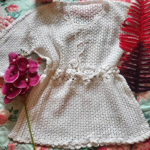 May include: A white crochet tunic featuring a V-neckline, three-quarter length sleeves, and a drawstring waist tie. The garment is crafted with an openwork stitch pattern and finished with a scalloped edge along the hem and sleeve cuffs.