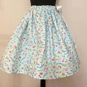 May include: A light blue skirt with a white ribbon tie. The skirt is printed with a whimsical pattern of narwhals, fish, and seaweed.