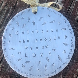 May include: A round, silver-colored ornament with the words "Celebrate the people you LOVE" stamped in the center. The ornament is decorated with stamped feather and heart designs around the edges and has a gold ribbon for hanging.