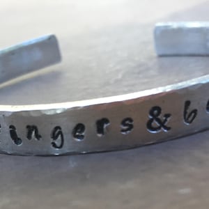 May include: A silver-toned cuff bracelet with the phrase "her fingers & bells" engraved in a handwritten style. The bracelet has a hammered texture and an open design.