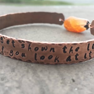 May include: Copper bracelet with a hammered finish and an orange gemstone charm. The bracelet has the inscription "You get shown the light if you look at it right".