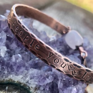 May include: Copper-toned cuff bracelet with the words "people you love" stamped on the front. The bracelet has a hammered texture and a small, light-colored stone accent. The bracelet is displayed on a purple crystal.