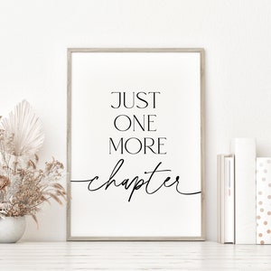 May include: A framed print with a white background and black text that reads "JUST ONE MORE chapter" in a cursive font. The print is framed in a light wood frame and is sitting on a white surface with dried flowers in a vase.