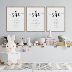 May include: Three framed prints with the word "she" in cursive script and bible verses about women. The prints are hung on a white wall above a white shelf with toys.