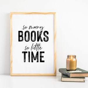 May include: A framed print with the text "So many BOOKS so little TIME" in black brush script on a white background. The print is sitting on a white surface next to a lit candle in a glass jar and a stack of books.