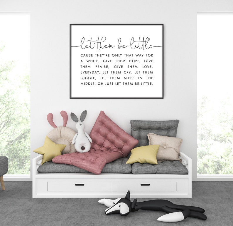 May include: A black framed print with white text that reads "let them be little" and a quote about letting children be children. The print is hanging on a white wall in a room with a white couch and pillows.