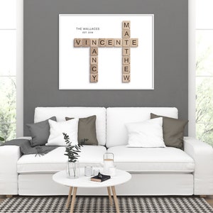 May include: A white framed print with a scrabble tile design spelling out the names "Vincent" and "Matthew" with the text "The Wallaces Est. 2018" at the top. The print is hanging on a gray wall above a white couch with gray and brown throw pillows.